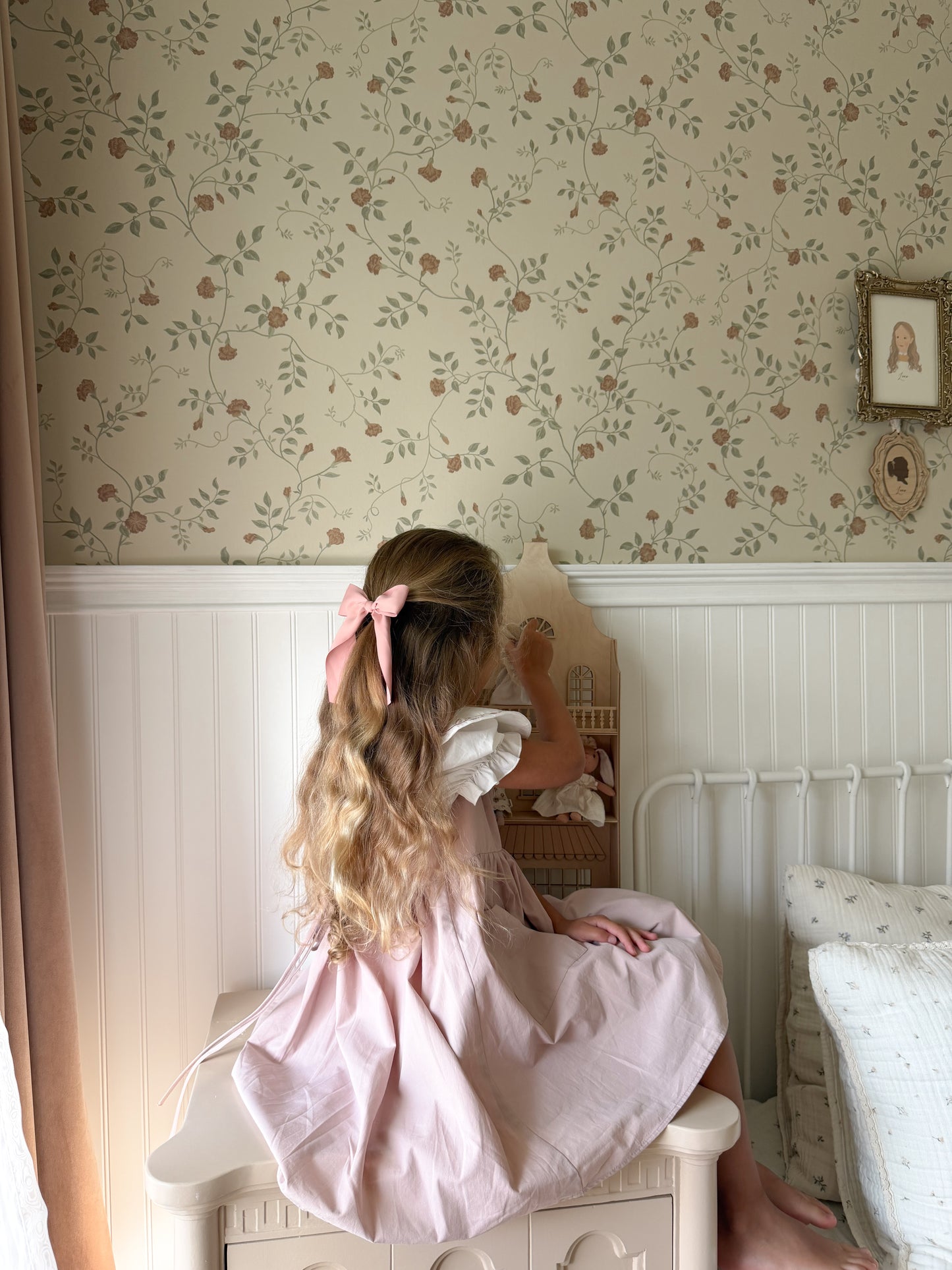 A young girl with long wavy blonde hair tied back with a pink bow sits on a white nightstand in a child's bedroom, looking at a small dollhouse. She is wearing a light pink dress.