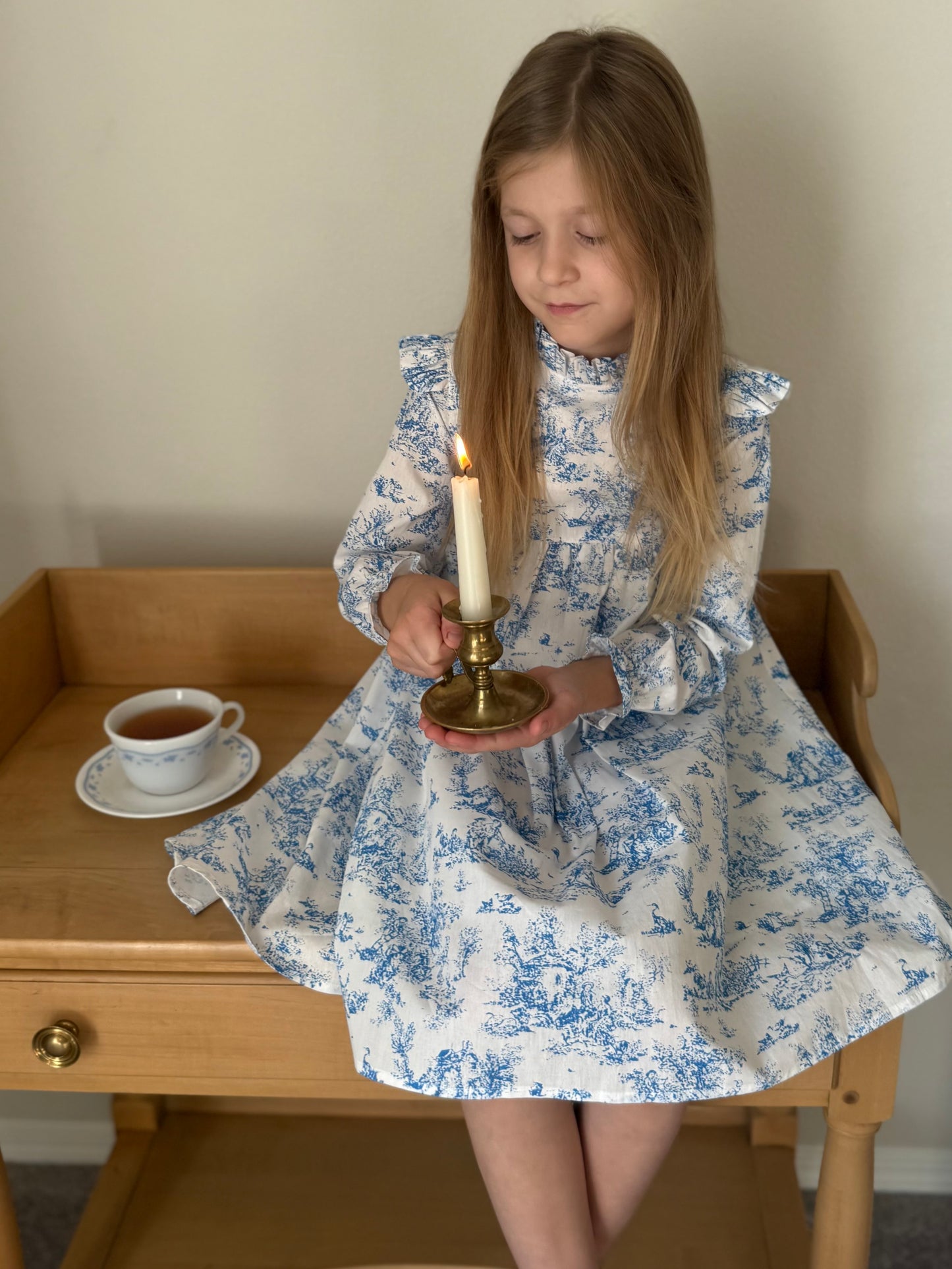 Young girl in a blue toile floral print dress holding a candle, sitting on a wooden table with a cup of tea.