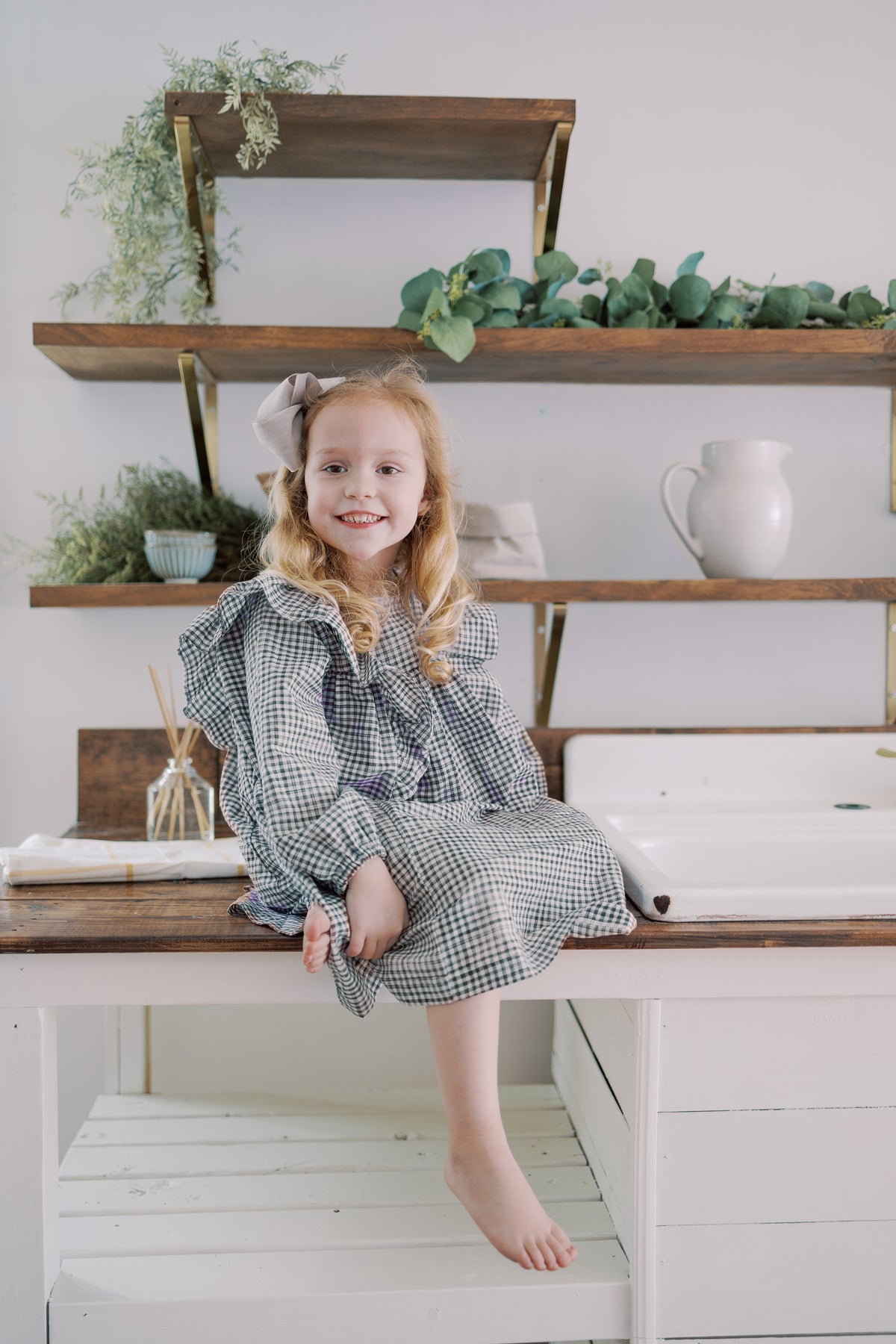A young girl with blonde hair sits barefoot on a white wooden table, wearing a long-sleeve gingham dress in a checkered gray and white pattern. She is posed in front of a rustic wall with floating wooden shelves holding green plants and a white ceramic pitcher.