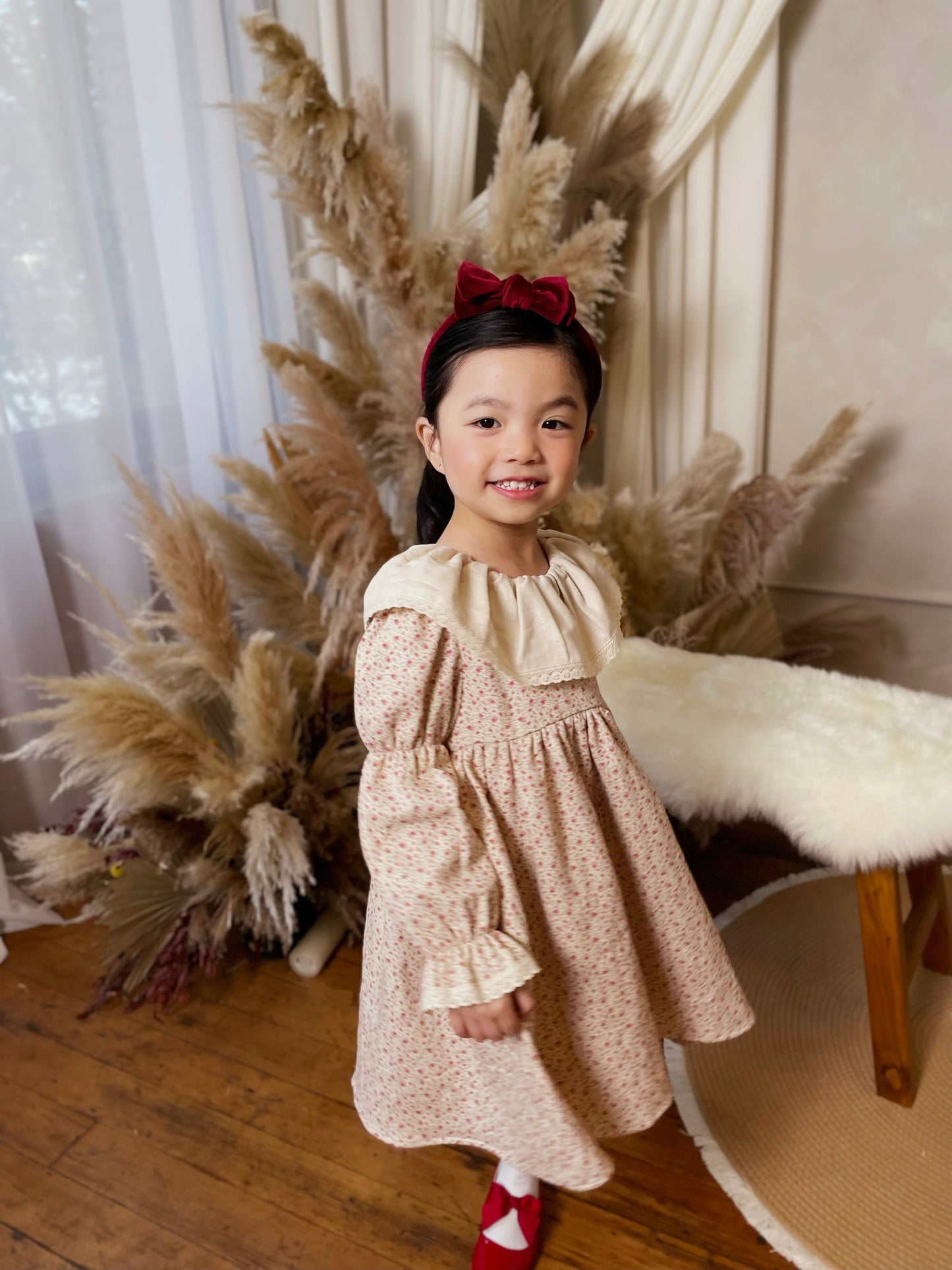 Young girl in a floral dress standing in front of decorative pampas grass and white curtains.
