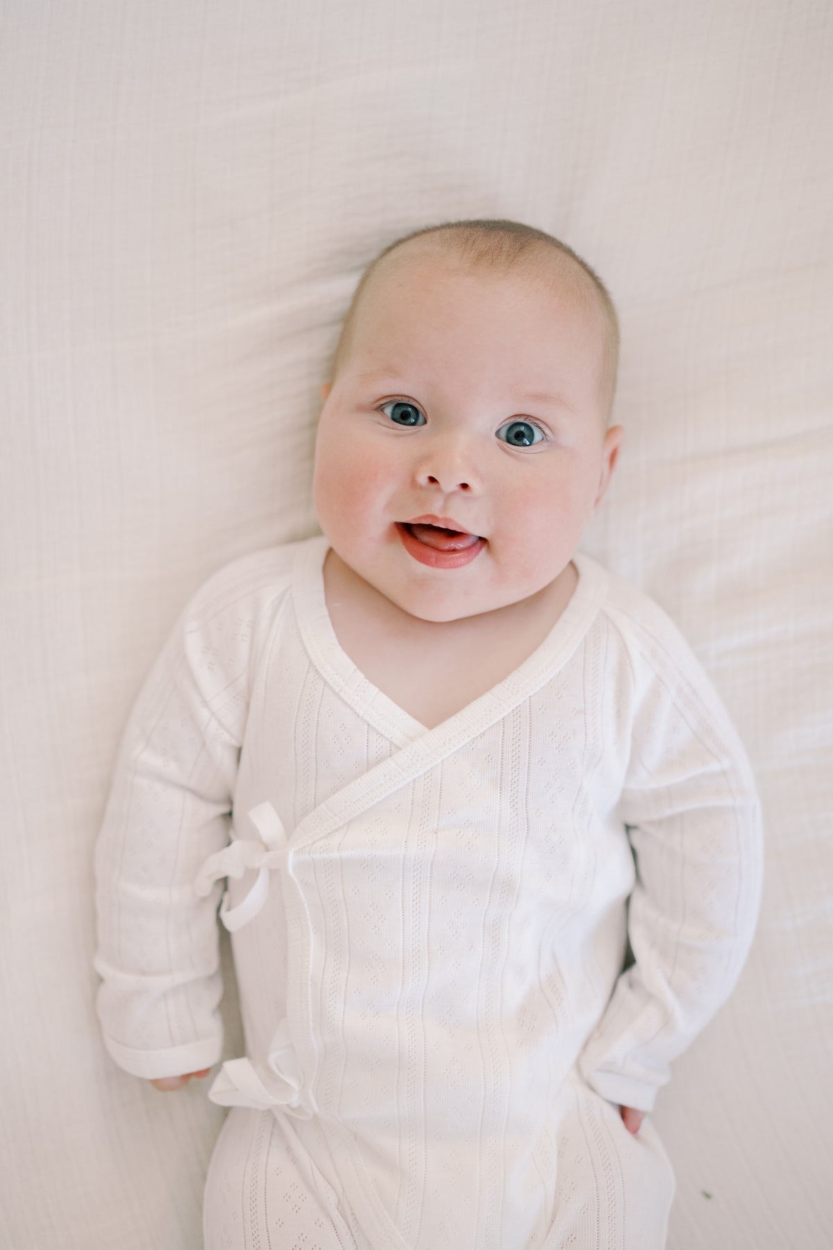 A smiling baby lying on a white textured blanket, wearing a white long-sleeve wrap bodysuit with a small bow detail on the chest.