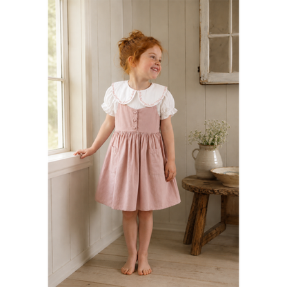 A young girl with red hair stands barefoot in a rustic room wearing a dusty pink pinafore dress with a white ruffled Peter Pan collar and white short sleeves. She is smiling and looking to the side near a window.