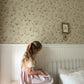 A young girl with blonde hair in a pink dress sits on a white wooden stool in a bedroom, reading a book. The room features floral wallpaper on the upper wall, white wainscoting, and a white metal bed frame with patterned bedding.