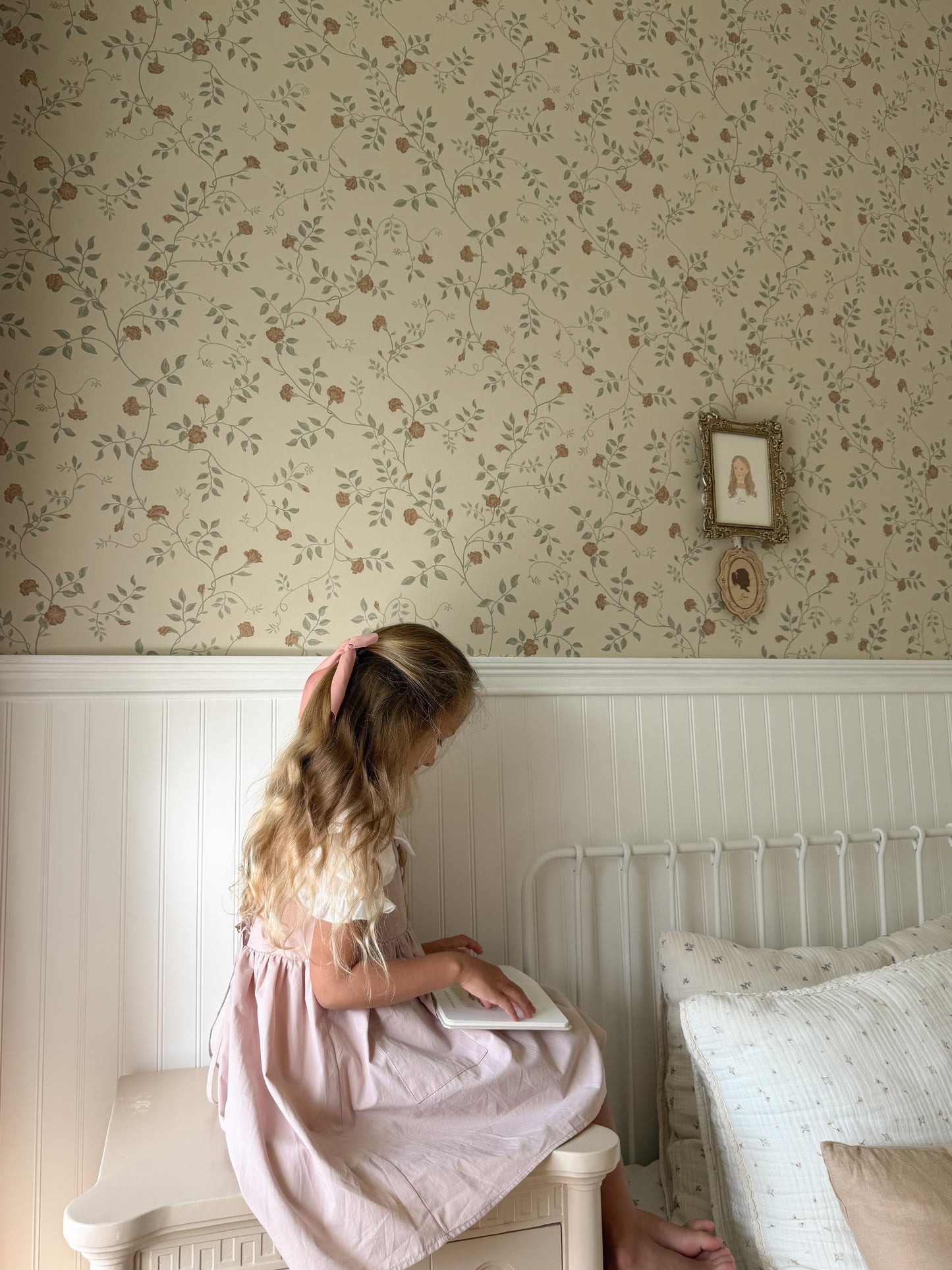 A young girl with blonde hair in a pink dress sits on a white wooden stool in a bedroom, reading a book. The room features floral wallpaper on the upper wall, white wainscoting, and a white metal bed frame with patterned bedding.