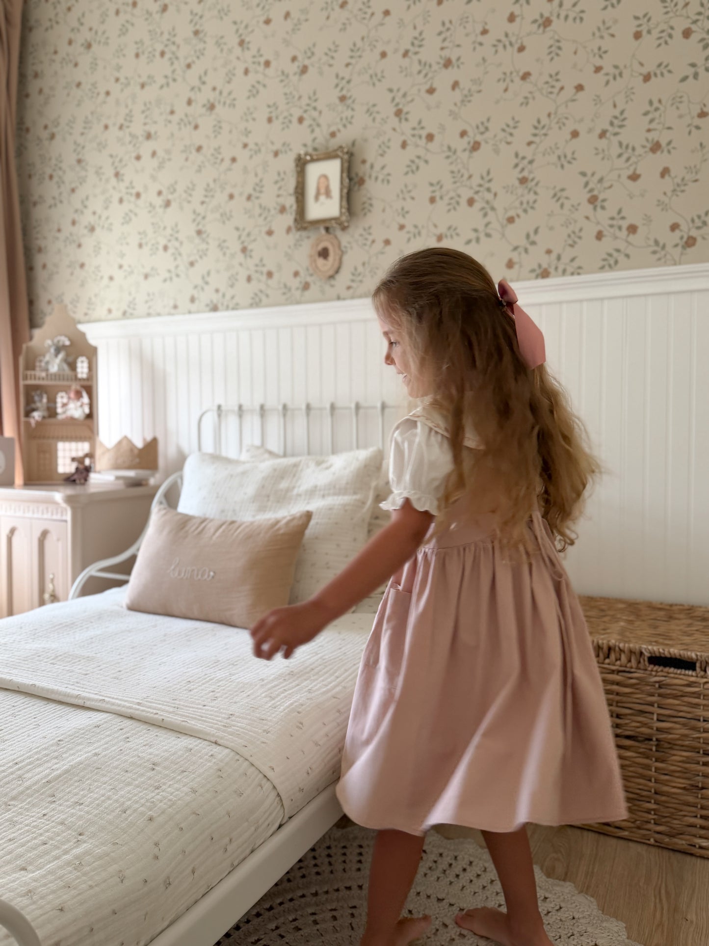 A young girl with long light brown hair tied back with a pink bow stands barefoot in a bedroom, wearing a dusty pink sleeveless pinafore dress over a white short-sleeve blouse. She is positioned next to a white bed with cream bedding and floral wallpaper in the background.