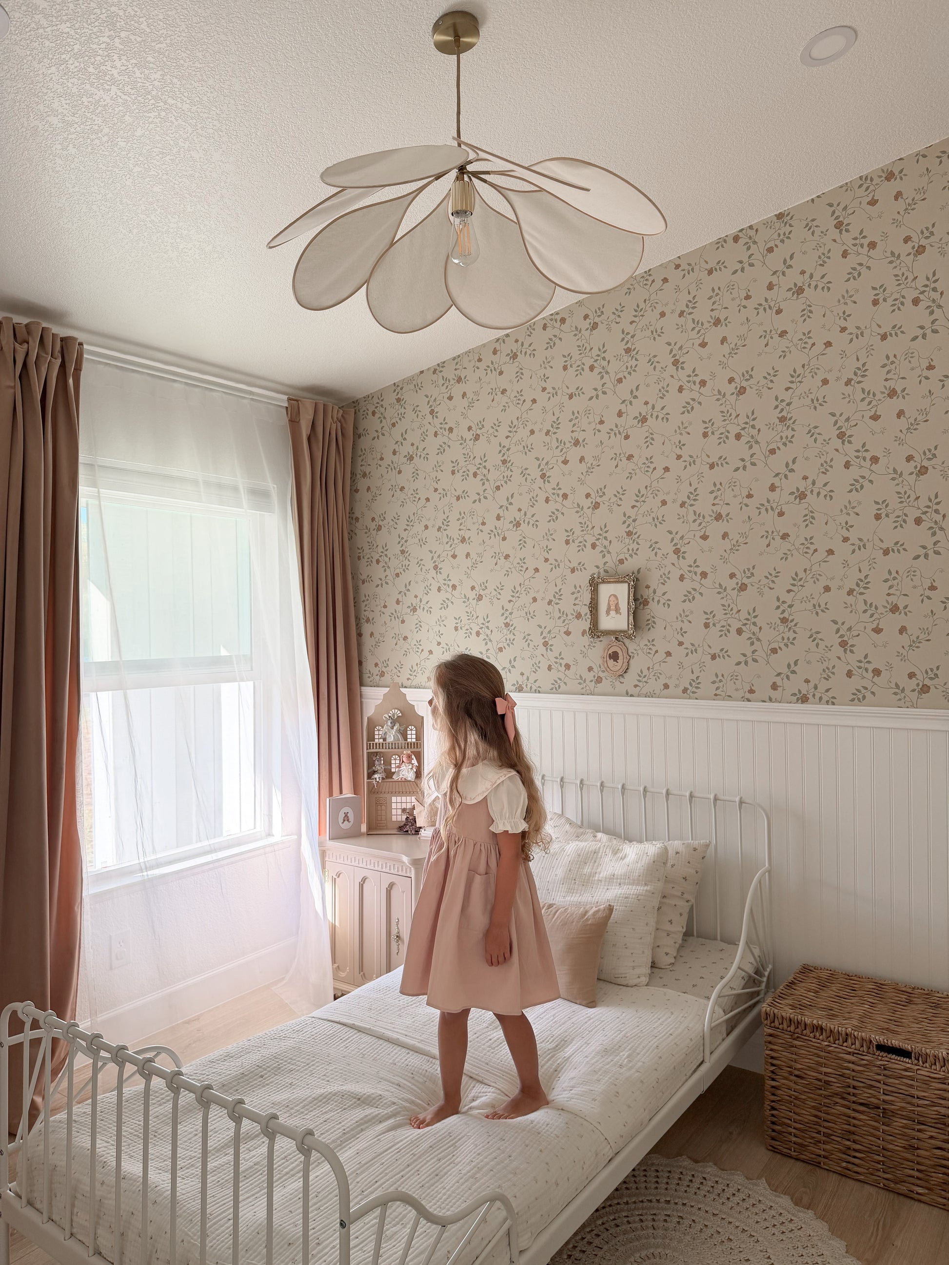 A young girl in a pink dress stands on a white metal bed in a softly lit bedroom with floral wallpaper, brown curtains, a flower-shaped ceiling light, and a wicker storage basket.