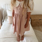 A young girl wearing a dusty pink pinafore dress with a white ruffled Peter Pan collar and short puff sleeves, standing barefoot on a white quilted bed in a bedroom.