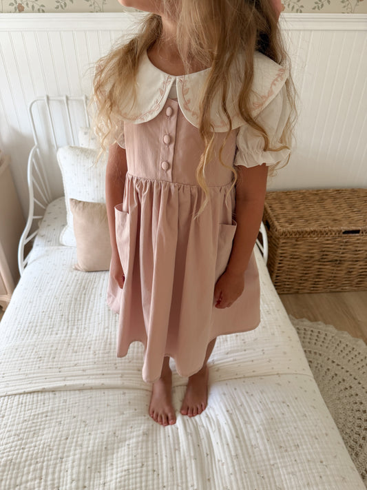 A young girl wearing a dusty pink pinafore dress with a white ruffled Peter Pan collar and short puff sleeves, standing barefoot on a white quilted bed in a bedroom.