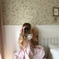 A young girl with long wavy blonde hair tied back with a pink bow sits on a white nightstand in a child's bedroom, looking at a small dollhouse. She is wearing a light pink dress.