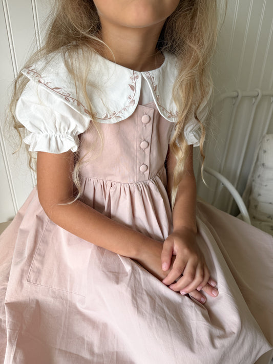 A young girl wearing a light pink dress with a white Peter Pan collar and ruffled sleeves, sitting indoors against a white paneled wall.