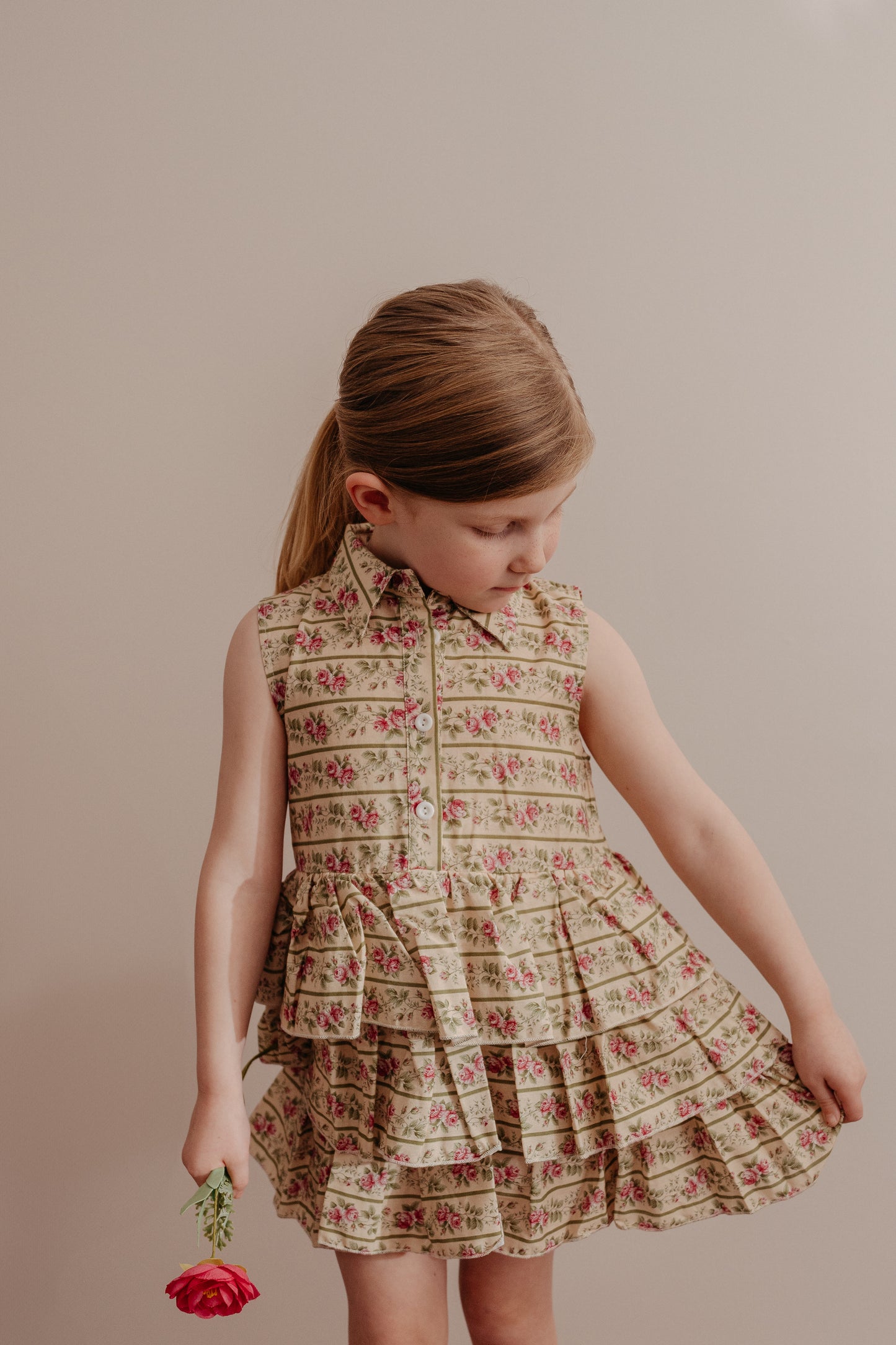 A young child wearing a sleeveless, floral print dress with a button-down front and a collar, featuring a multi-tiered ruffle skirt.