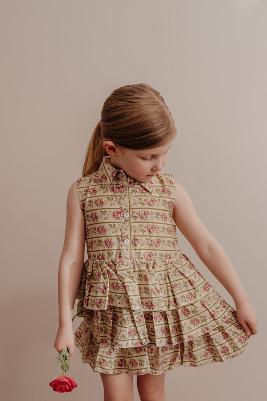 A young child wearing a sleeveless, floral print dress with a button-down front and a collar, featuring a multi-tiered ruffle skirt.