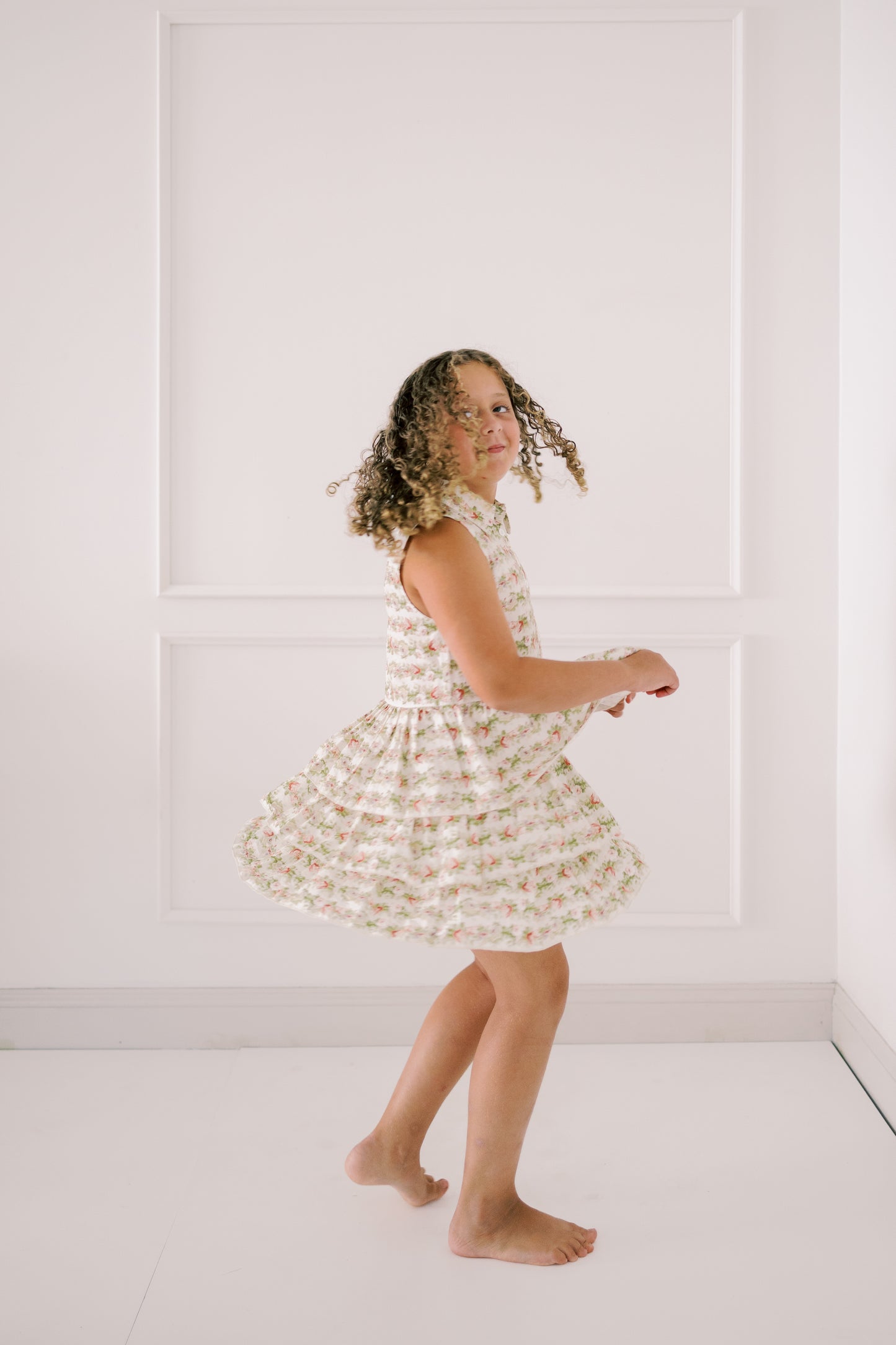 Young girl in a floral dress standing against a white paneled wall.