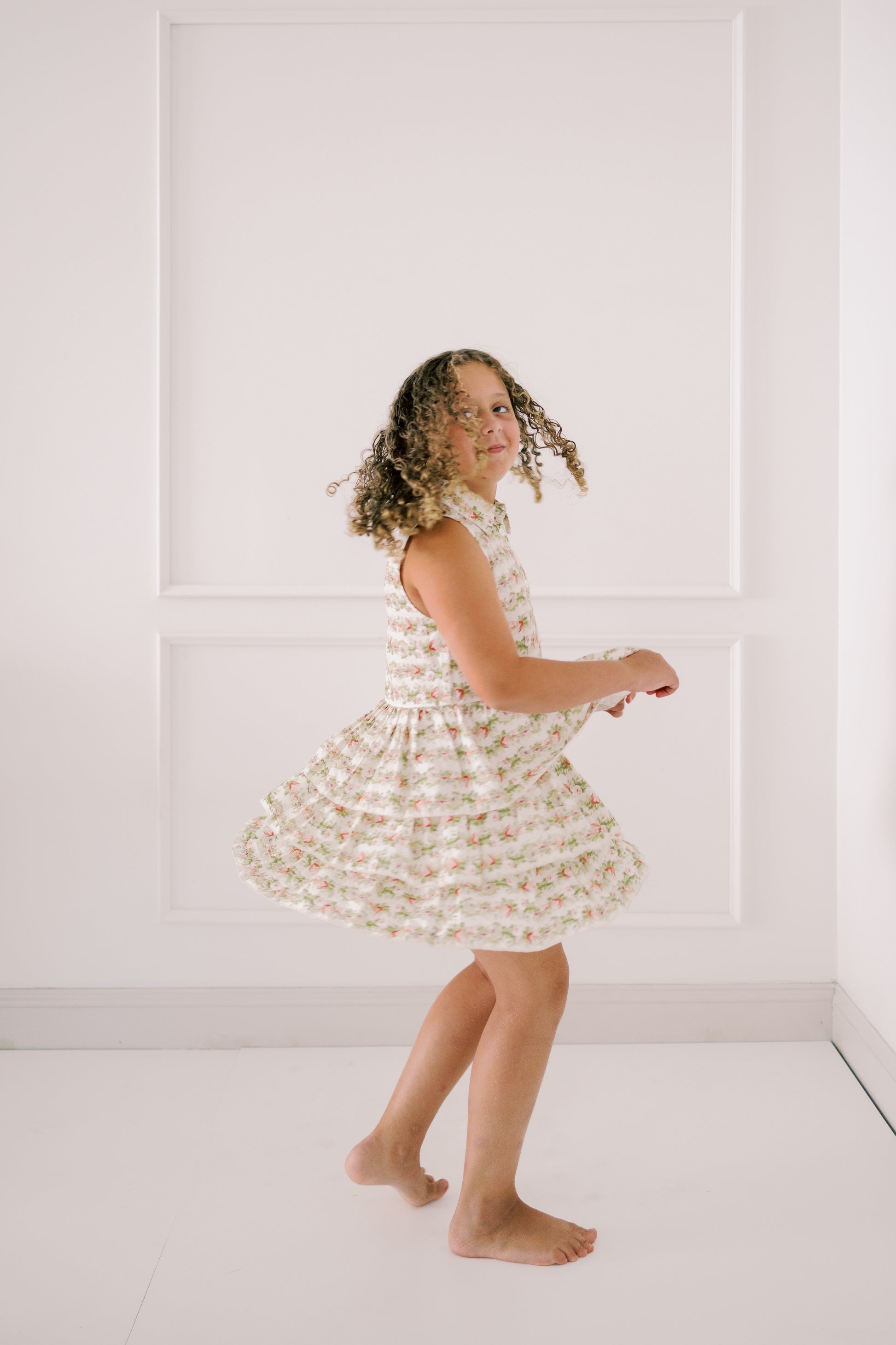 Young girl in a floral dress standing against a white paneled wall.