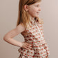 A young girl wearing a sleeveless, floral print, button-down, multi-tiered ruffle skirt dress.
