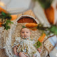 Baby in a floral dress sitting on a textured surface with pumpkins and greenery in the background