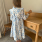 Child wearing a blue floral dress standing next to a wooden desk.