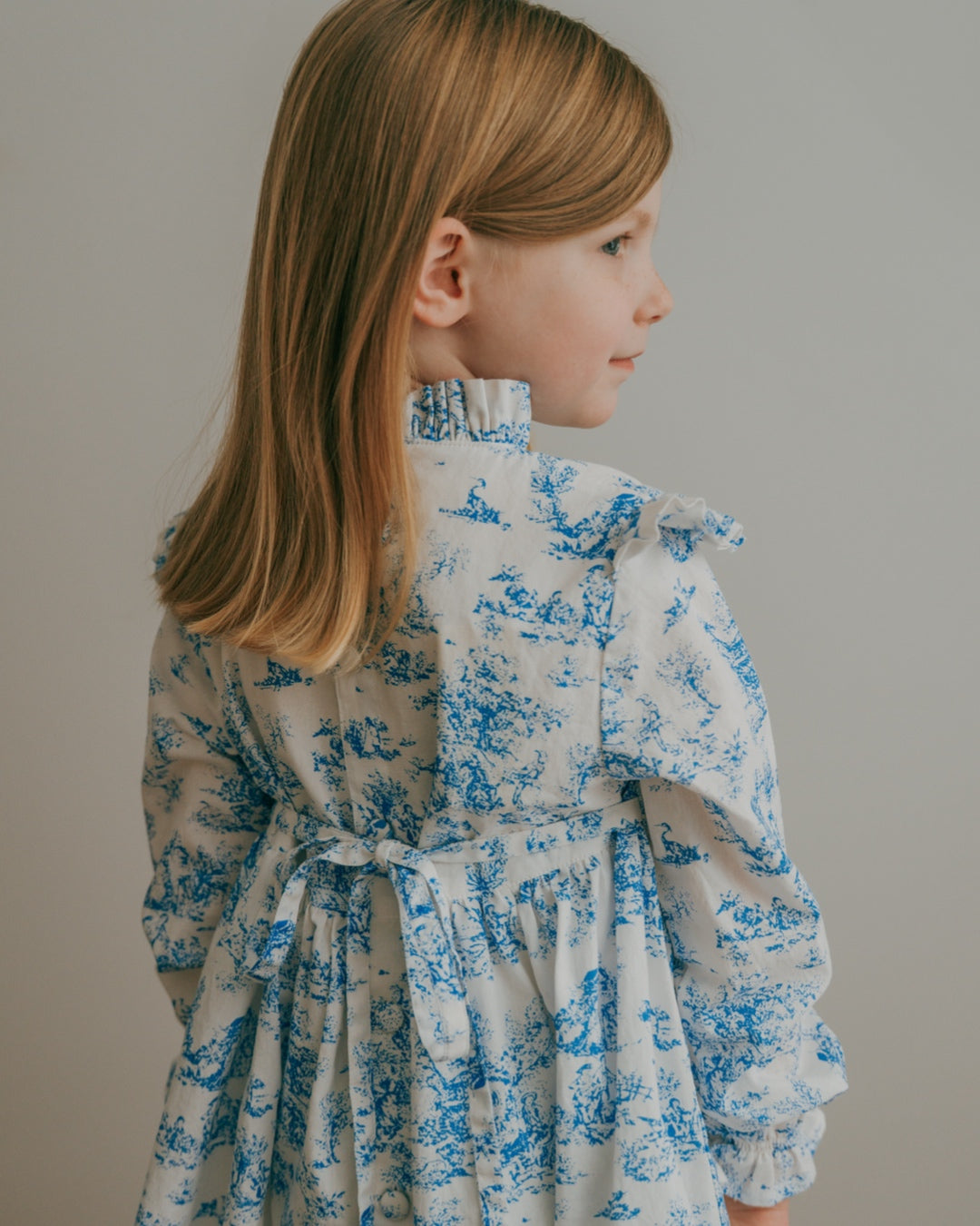 Young girl wearing a blue toile floral dress against a plain background