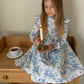 Young girl in a blue toile floral print  dress holding a candle, sitting on a wooden table with a cup of tea.