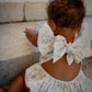 Child wearing Confetti Print Josie Bubble back view with large bows sitting on stone steps.