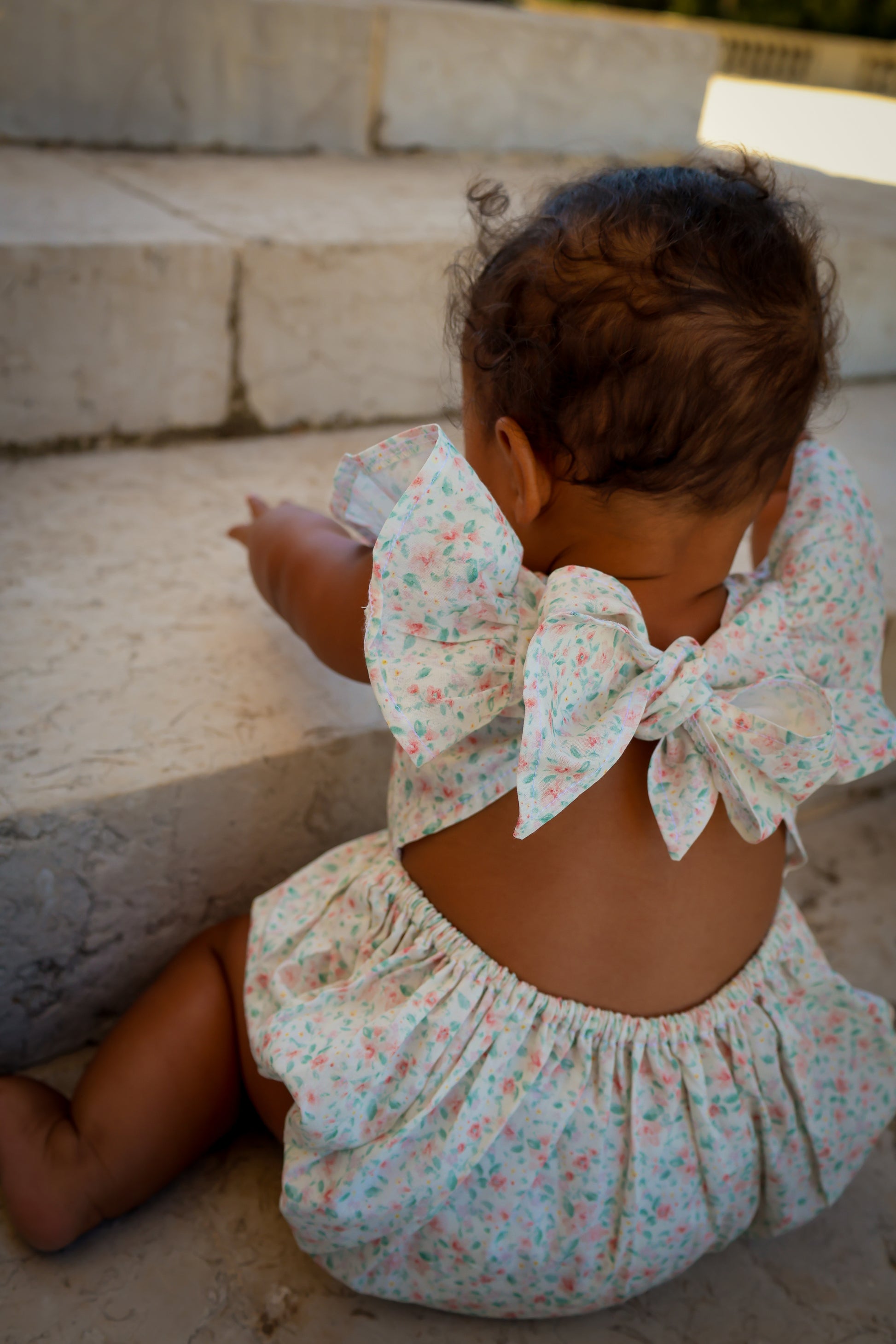 Child wearing Confetti Print Josie Bubble back view with large bows sitting on stone steps.