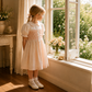 A young girl in a light pink and white dress stands by a window in a sunlit room, looking out onto a garden. A matching floral side table with flowers is visible beside her.