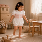 A toddler girl walking barefoot in a sunlit nursery, wearing a white short-sleeve romper with pink smocked detailing across the chest.