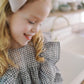 A young girl with blonde hair wearing a purple and white gingham ruffled top and a large light gray bow headband, smiling in a bright kitchen setting.