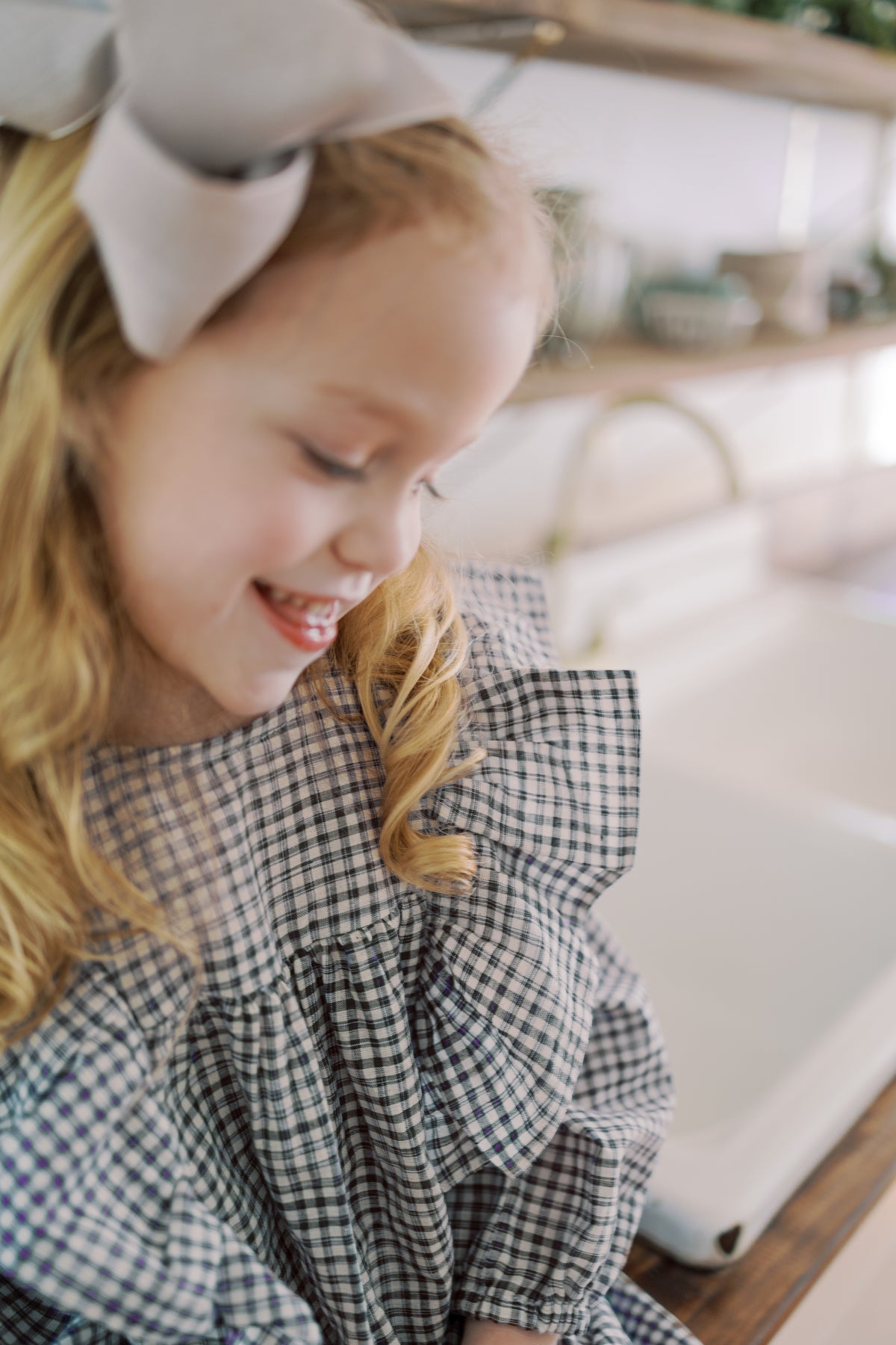 A young girl with blonde hair wearing a purple and white gingham ruffled top and a large light gray bow headband, smiling in a bright kitchen setting.