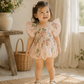 A toddler girl wearing a light pink floral romper dress with flutter sleeves, standing on a woven rug in a bright, sunlit room.