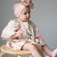 Baby sitting on a wicker chair wearing a floral bubble and pink headband with a bow.