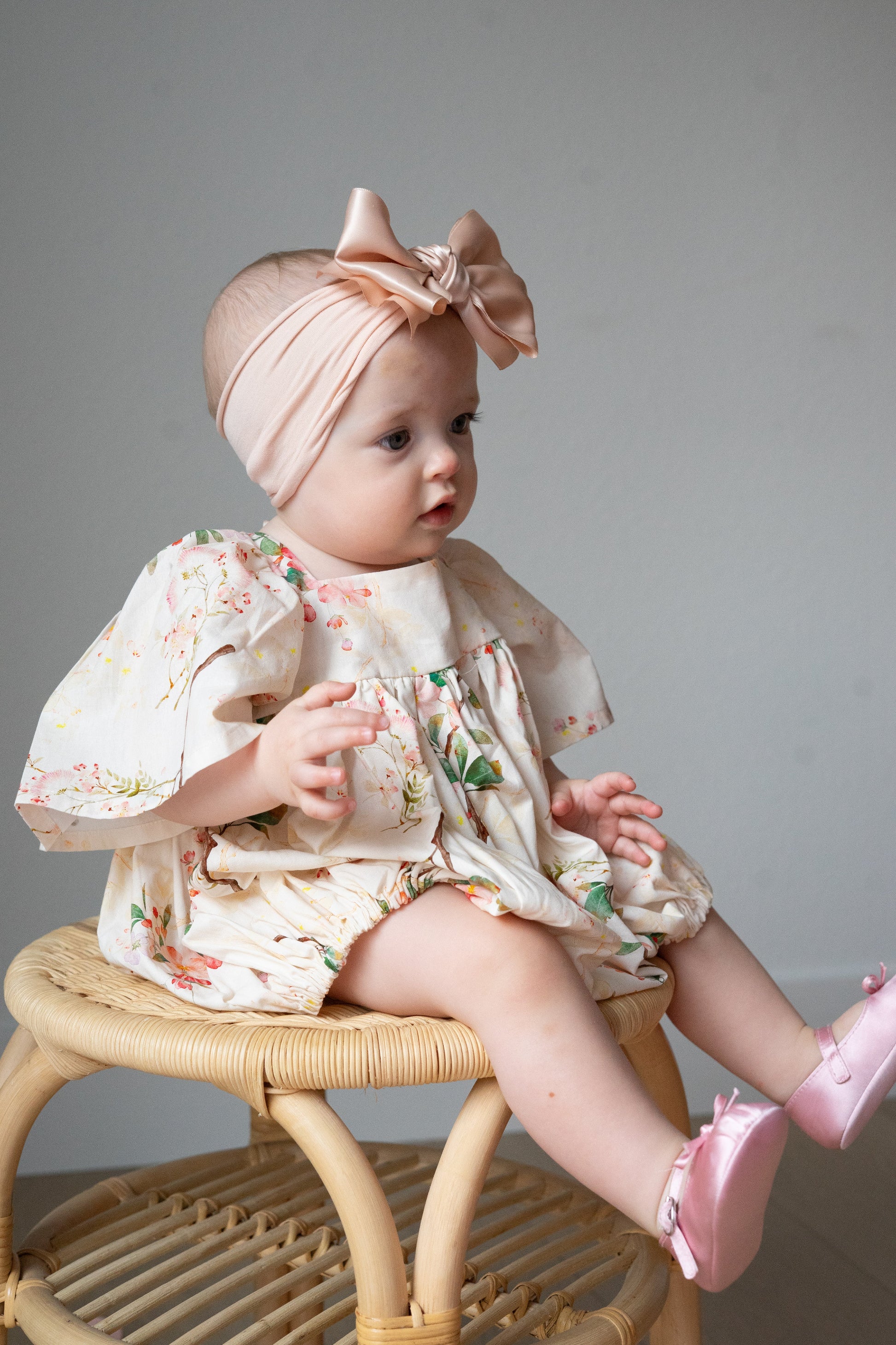 Baby sitting on a wicker chair wearing a floral bubble and pink headband with a bow.