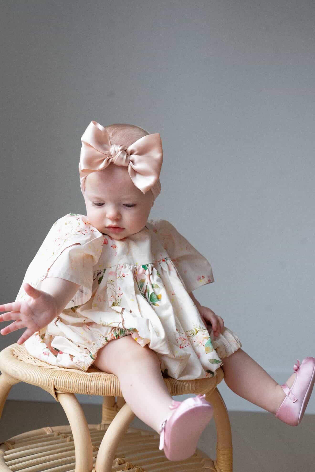 Baby sitting on a small stool wearing a floral dress and pink shoes with a large bow headband.