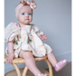 Baby sitting on a chair wearing a floral dress and pink shoes with a bow headband.