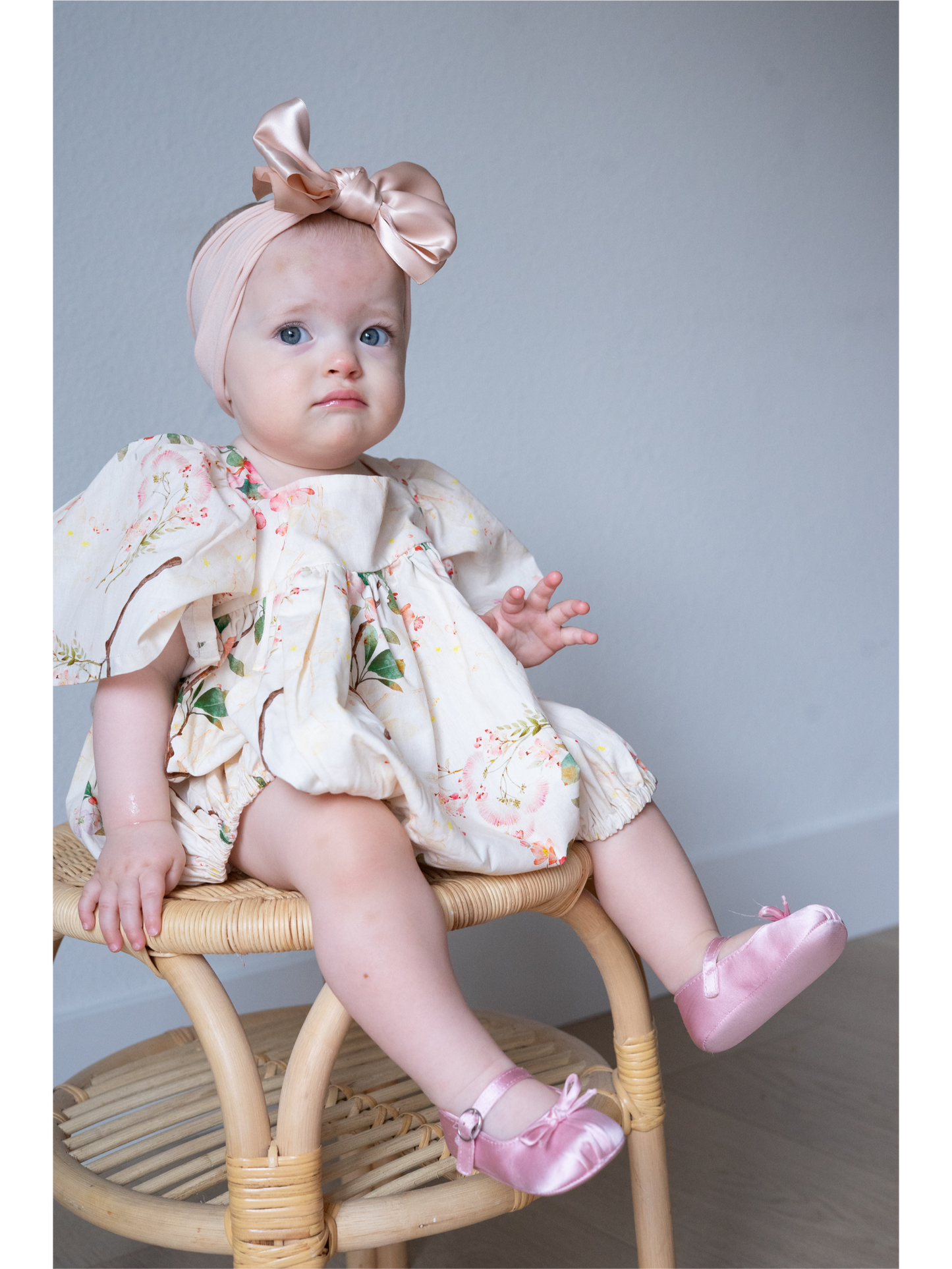 Baby sitting on a chair wearing a floral dress and pink shoes with a bow headband.