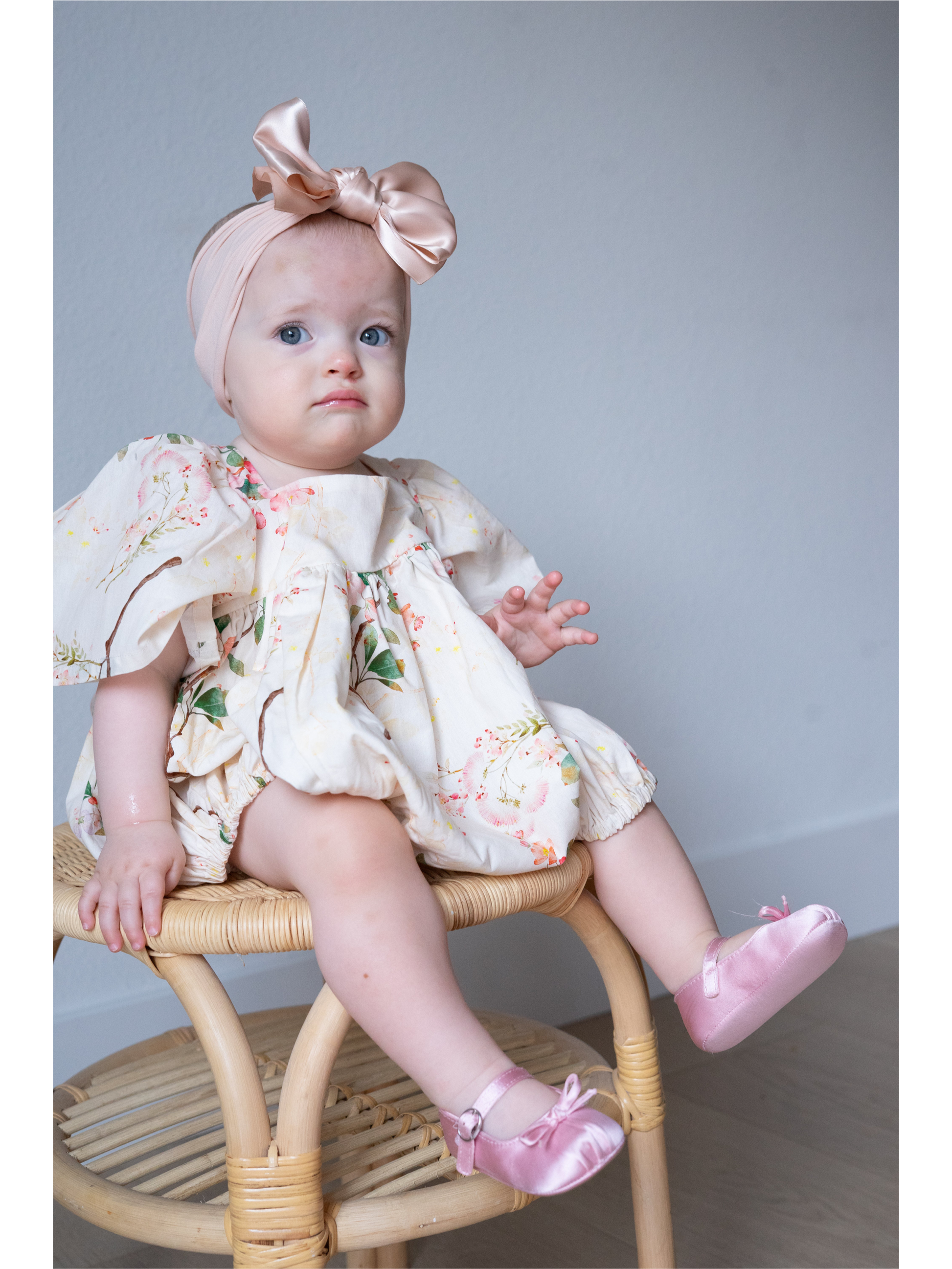 Baby sitting on a chair wearing a floral dress and pink shoes with a bow headband.