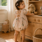 A toddler girl stands barefoot in a sunlit nursery or playroom, wearing a light blush pink floral romper with lace-up back ties. The room has warm wood cabinetry, baskets, a stuffed bunny, and a window with soft natural light.