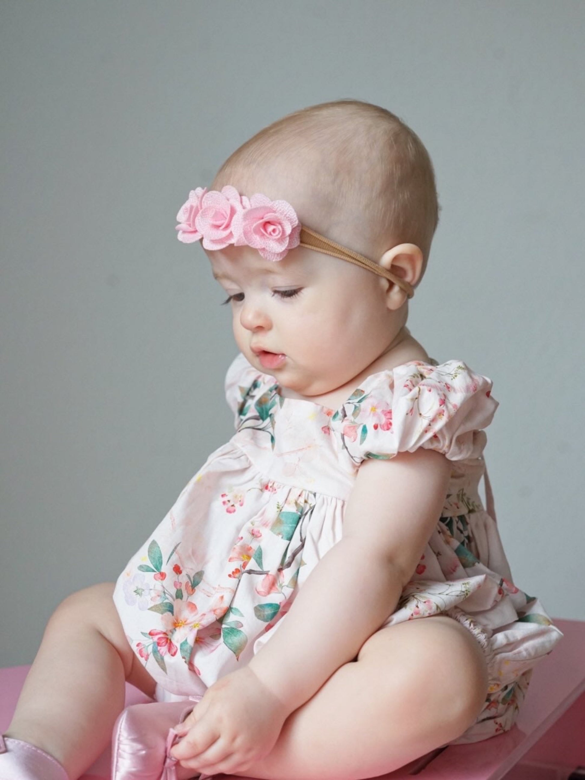 Baby wearing a floral Bubble and headband sitting on a pink surface with a gray background
