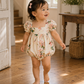 A toddler girl walking barefoot on a wooden floor in a bright room, wearing a cream puff-sleeve romper with a pink and green floral print and a small pink hair bow.