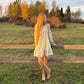 Woman with long yellow hair standing near a wooden fence in a grassy field with trees in the background.