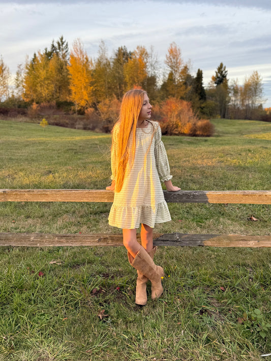 Woman with long yellow hair standing near a wooden fence in a grassy field with trees in the background.
