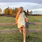 Young girl in a white dress and tan boots sitting on a wooden fence in a field.