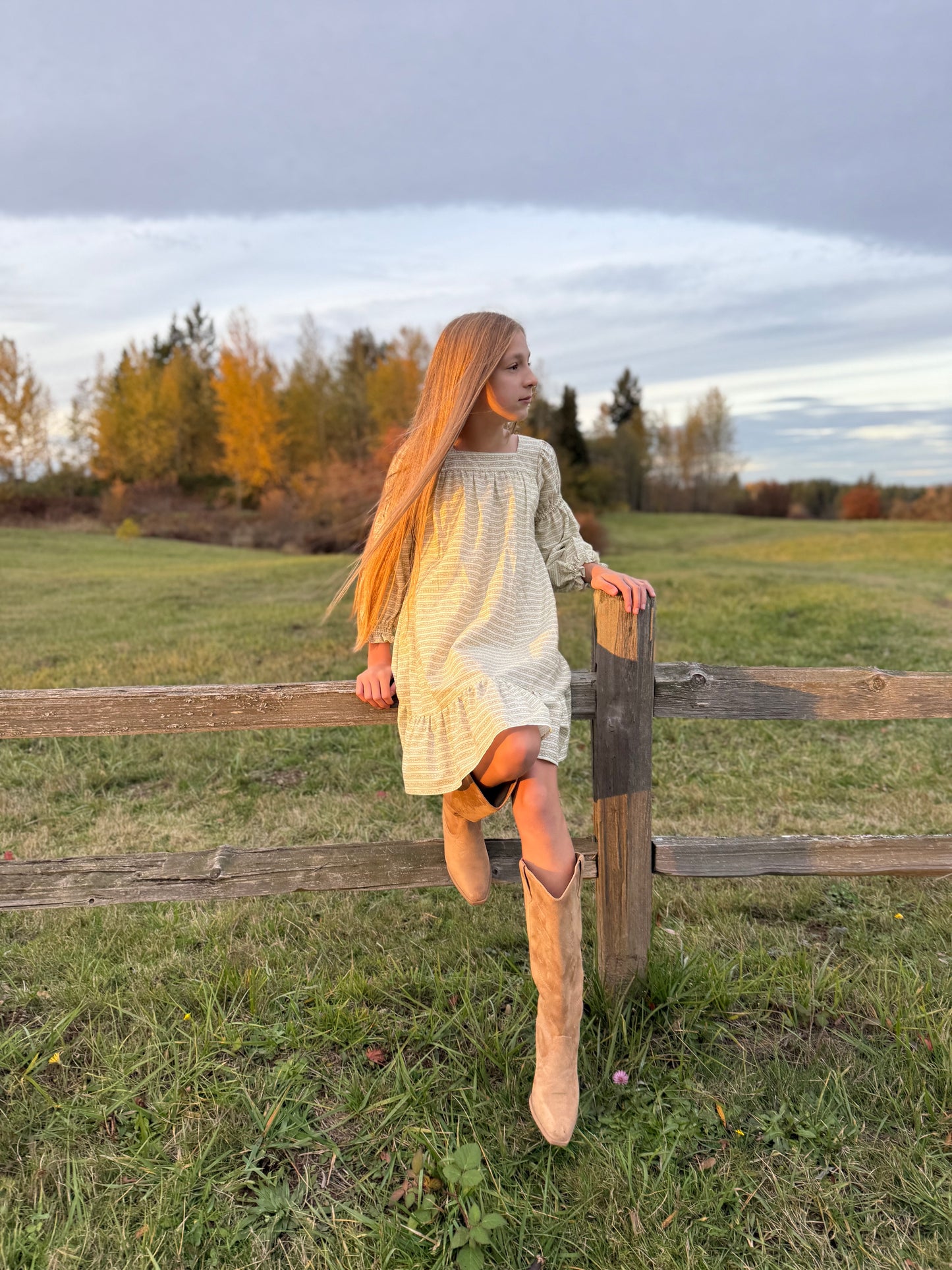 Young girl in a white dress and tan boots sitting on a wooden fence in a field.