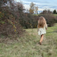 Person with long hair walking through a grassy field with trees and a blue sky in the background