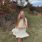 Young girl in a white dress and brown boots standing in a grassy field with trees in the background.