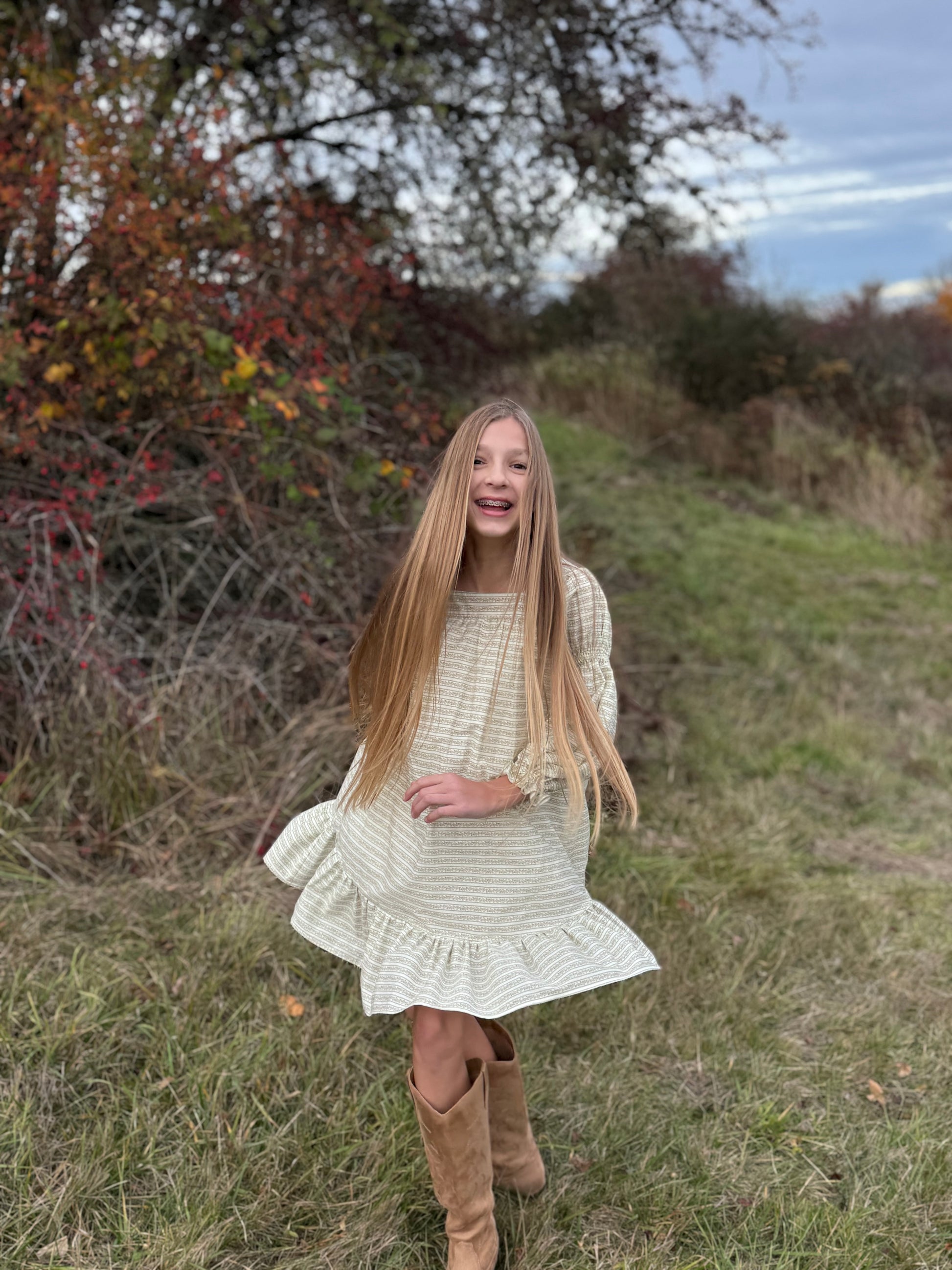Young girl in a white dress and brown boots standing in a grassy field with trees in the background.