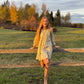 Young girl in a dress standing on a wooden fence in a grassy field with trees in the background.