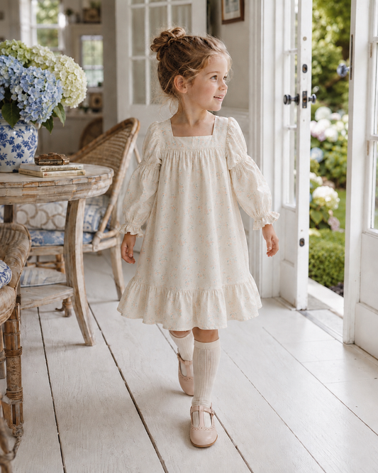 A young girl with her hair in a bun walks through a bright, white-floored interior doorway, wearing a cream-colored long-sleeve dress with a subtle floral print, matching cream knee-high socks, and light pink shoes.