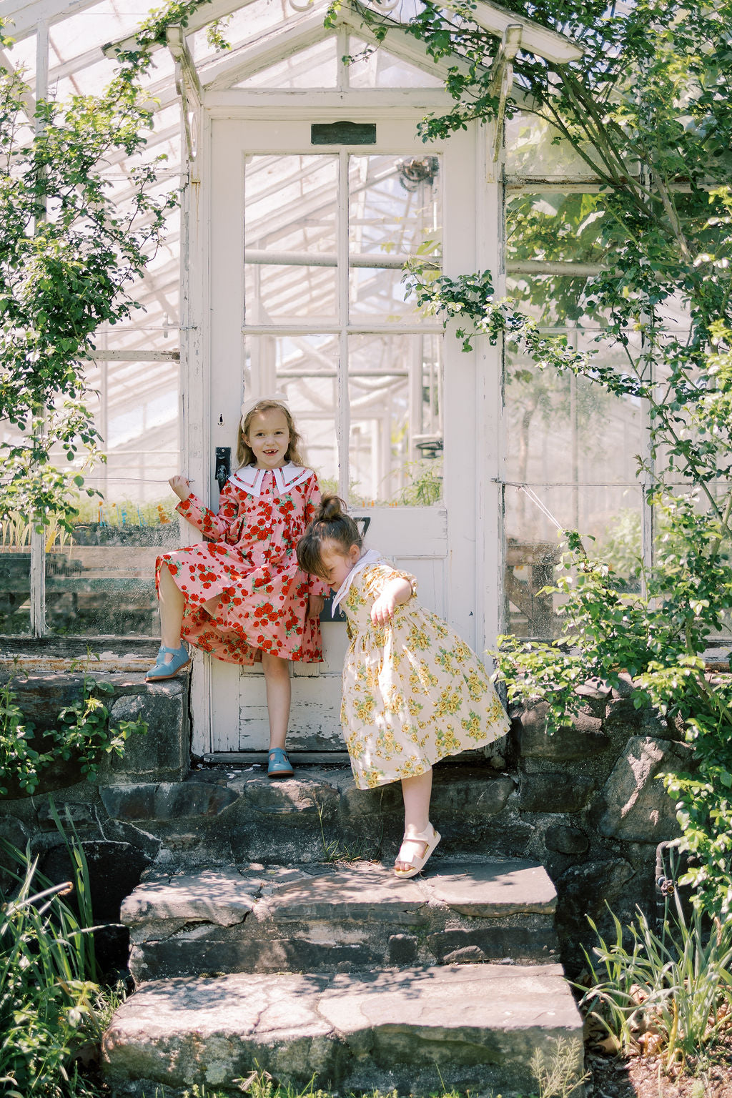Two children in colorful dresses standing on stone steps in a greenhouse.