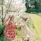 Two young girls in floral dresses standing in a garden with cherry blossom trees.